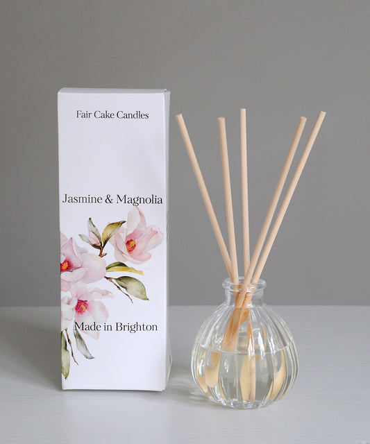Diffuser with wooden sticks and packaging labeled 'Fair Cake Candles Jasmine & Magnolia Made in Brighton' on a gray background.