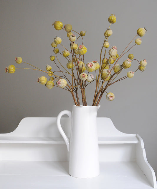 White vase with faux poppy seed head stems on a white surface against a gray background