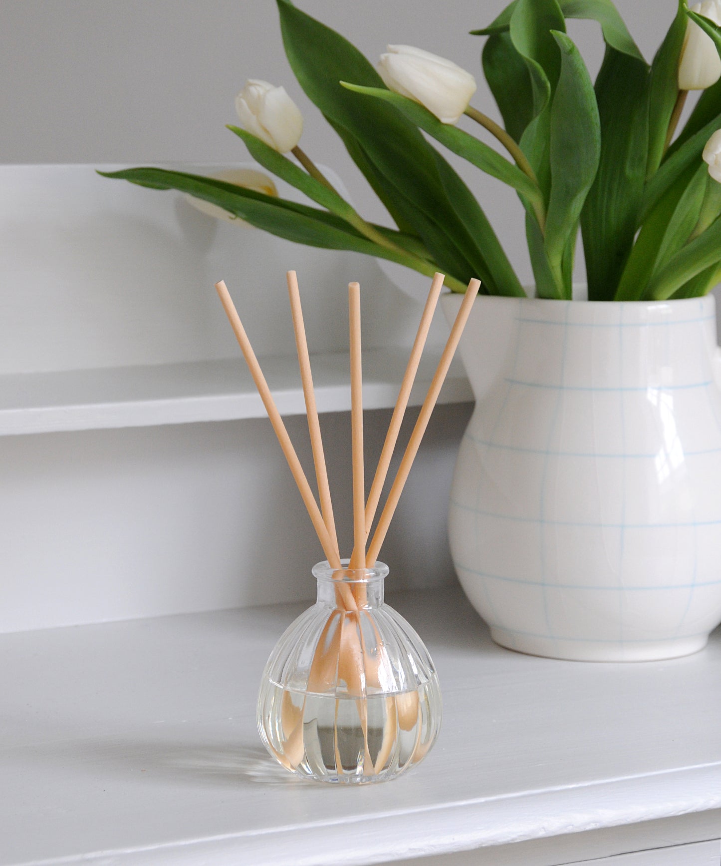 Clear diffuser bottle with wooden sticks next to a white vase with tulips on a white surface