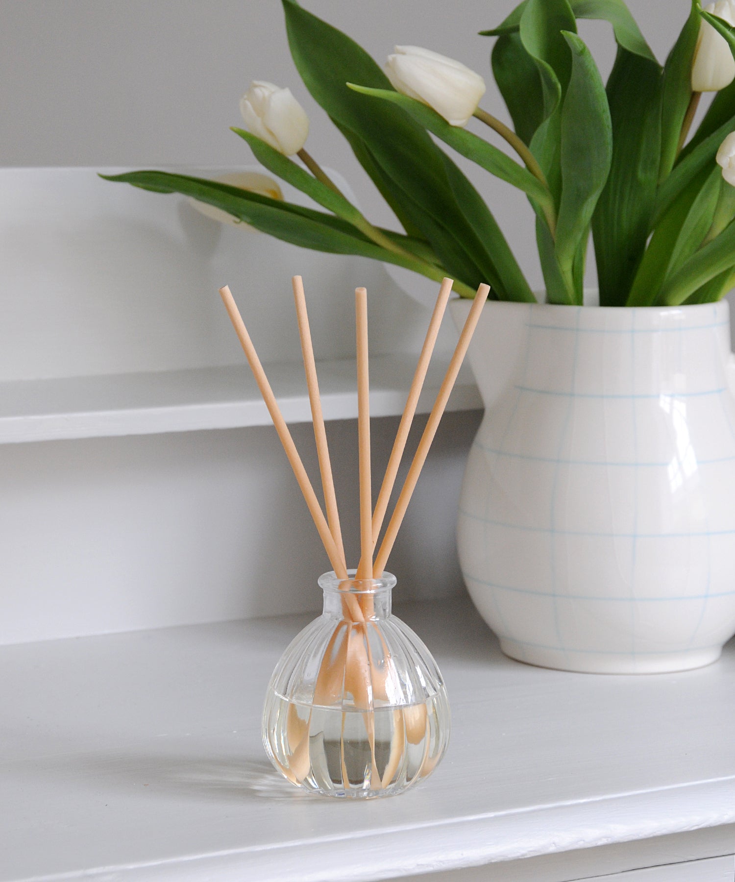 Clear diffuser bottle with wooden sticks next to a white vase with tulips on a white surface
