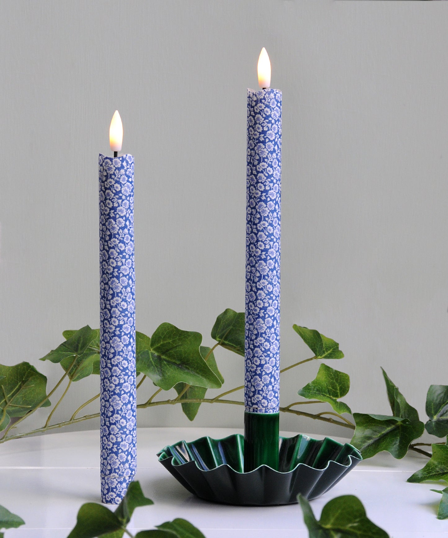 Two blue patterned candles in a green holder with leaves in the background
