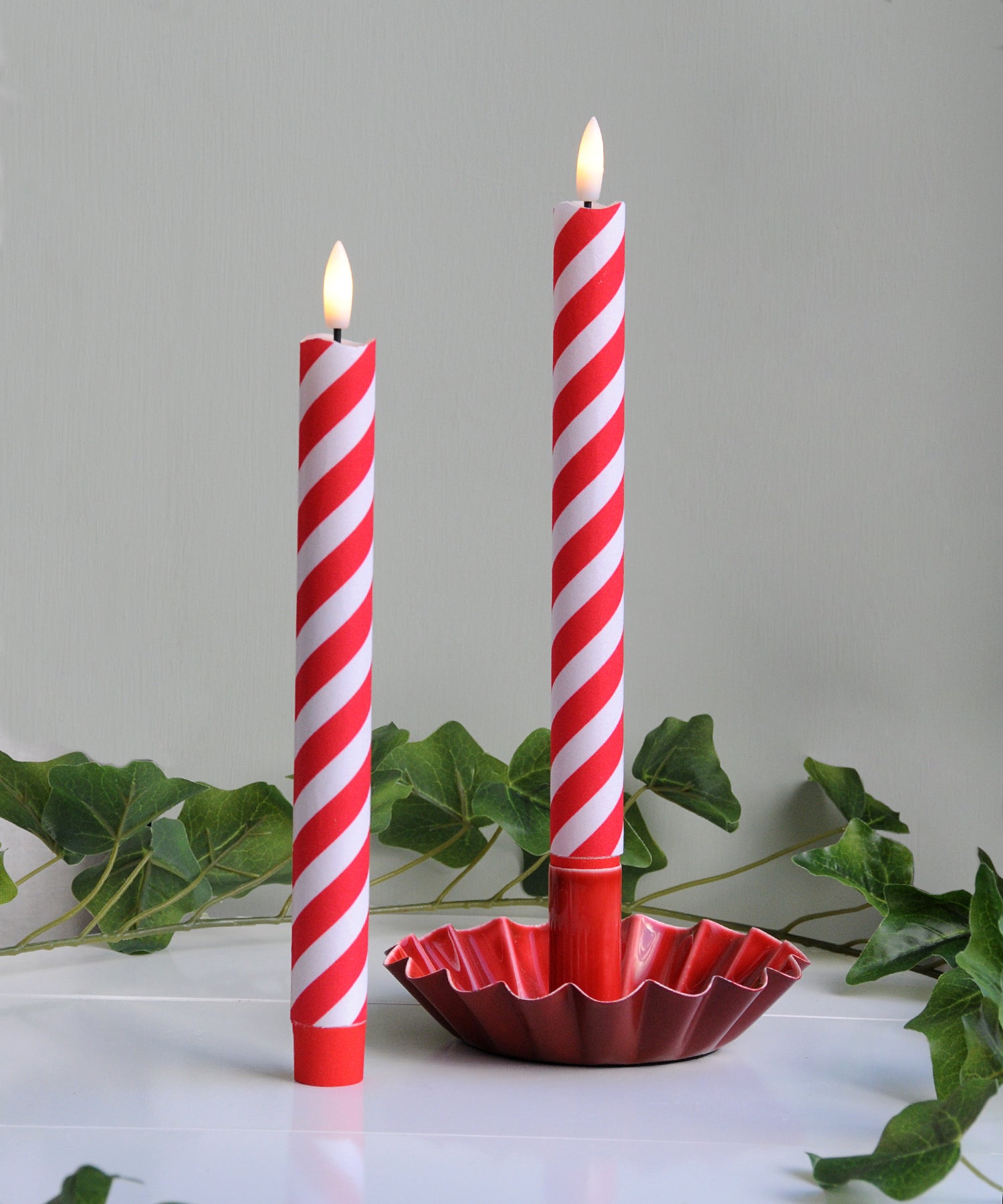 Two red and white striped candles in a red holder with green leaves in the background.